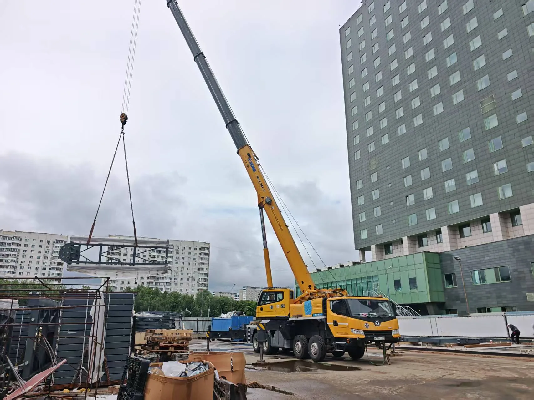 XCMG mobile crane lifting a steel parking platform module on the Moscow automated parking installation site, with high-rise residential towers in the background